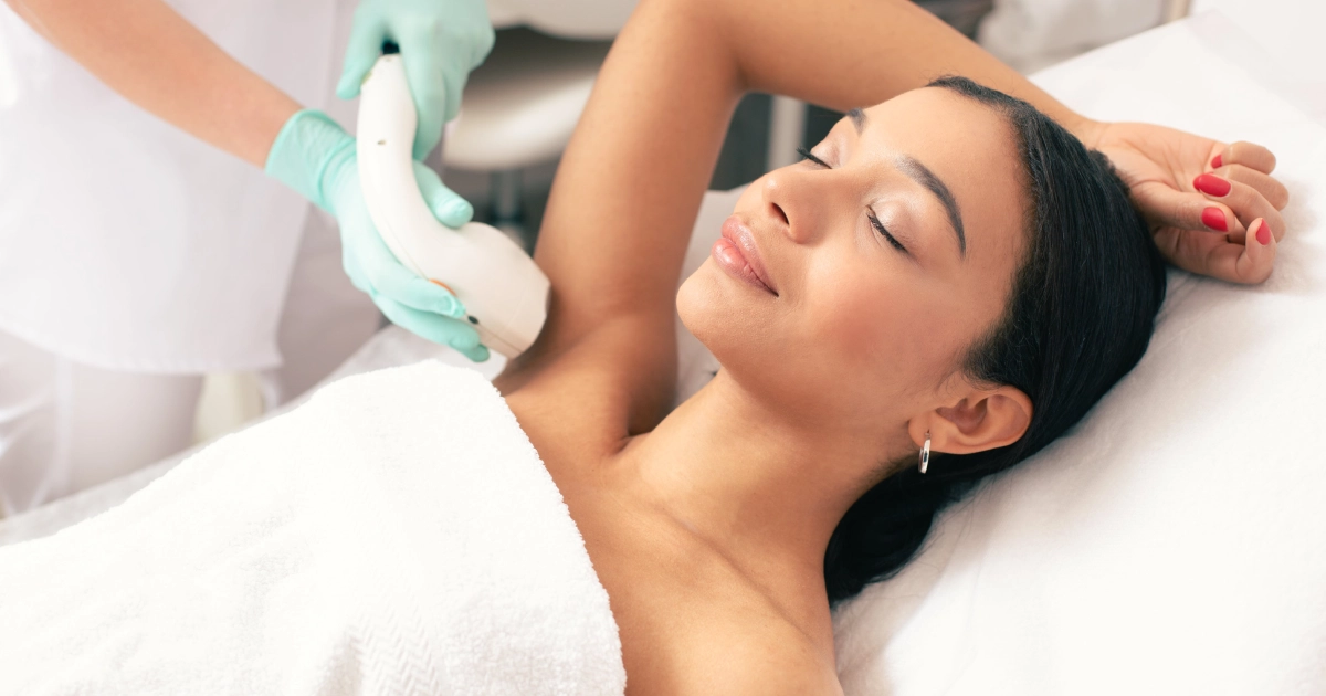 A woman relaxing on a treatment bed while a professional technician performs laser hair removal in Fredericksburg, VA on her underarm using a handheld device.